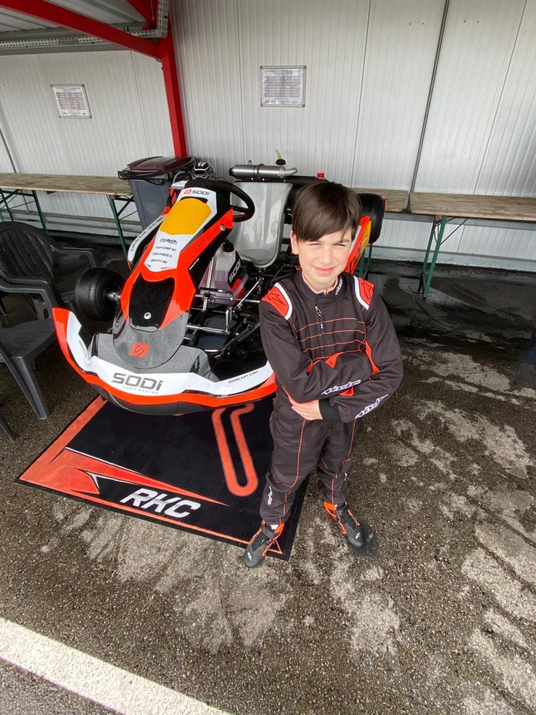 Le jeune pilote en combinaison à côté d’un kart au garage.